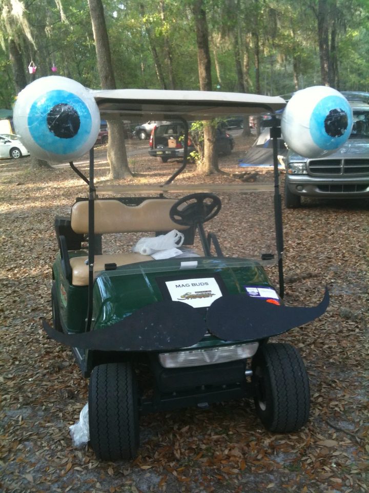 The original Zerble at a wooded campsite in 2011: a green golf cart with giant blue eyeballs mounted on the roof corners, a black posterboard mustache, and a MAG BUDS sign on the hood.