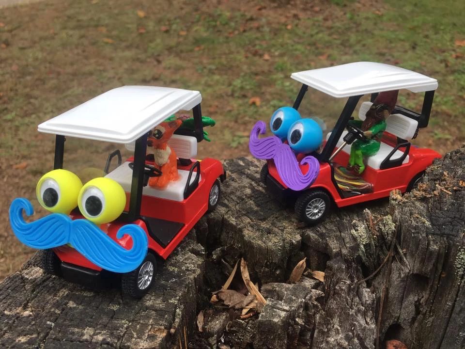 Two toy golf carts on a tree stump, each fitted with googly eyes and a curly mustache, one blue and one purple.