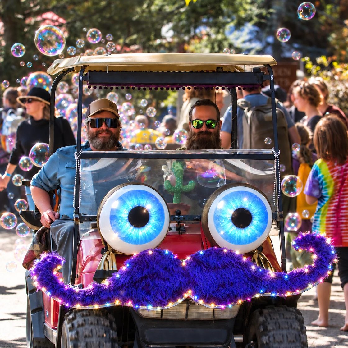 Gary and Locke drive Zerble through a festival crowd on a sunny day: a red golf cart with giant blue-and-white eyes, a purple fur mustache trimmed in lights, and bubbles floating all around.