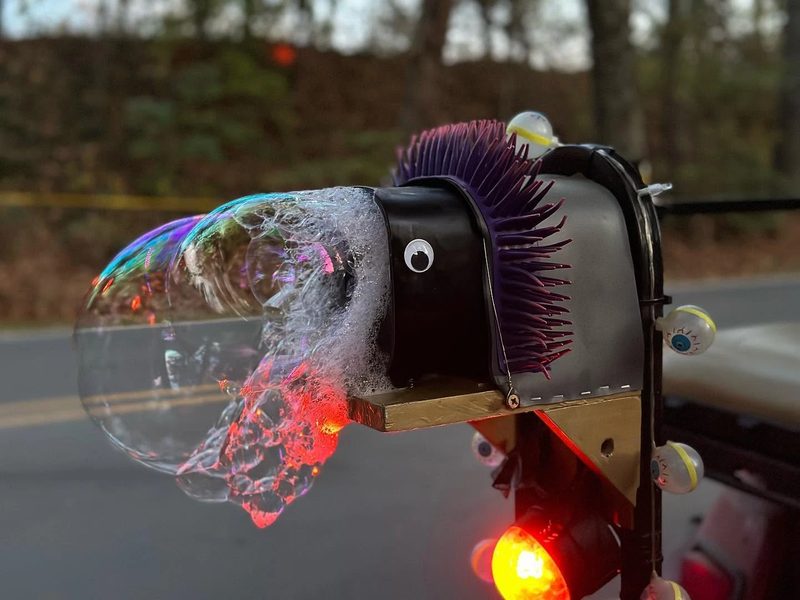 Close-up of one of Zerble's bubble machines mounted on the cart: a black housing with a googly eye and a purple spiky mohawk, blowing a big iridescent bubble.