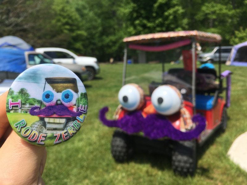 A round pin-back button reading I RODE ZERBLE, held up in front of the real Zerble: a red golf cart with giant white eyeballs and a fuzzy purple mustache, parked at a festival campsite.