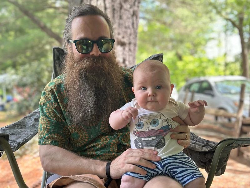 Baby Darwin in a hand-drawn Zerble onesie, sitting on a lap at a sunny campsite.