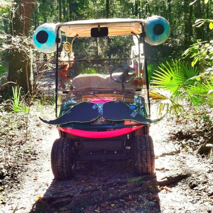 An early Zerble on a wooded festival trail: a golf cart with blue eyeballs on the roof corners, a black posterboard mustache outlined in glowing EL wire, and a pink pool-noodle smile.