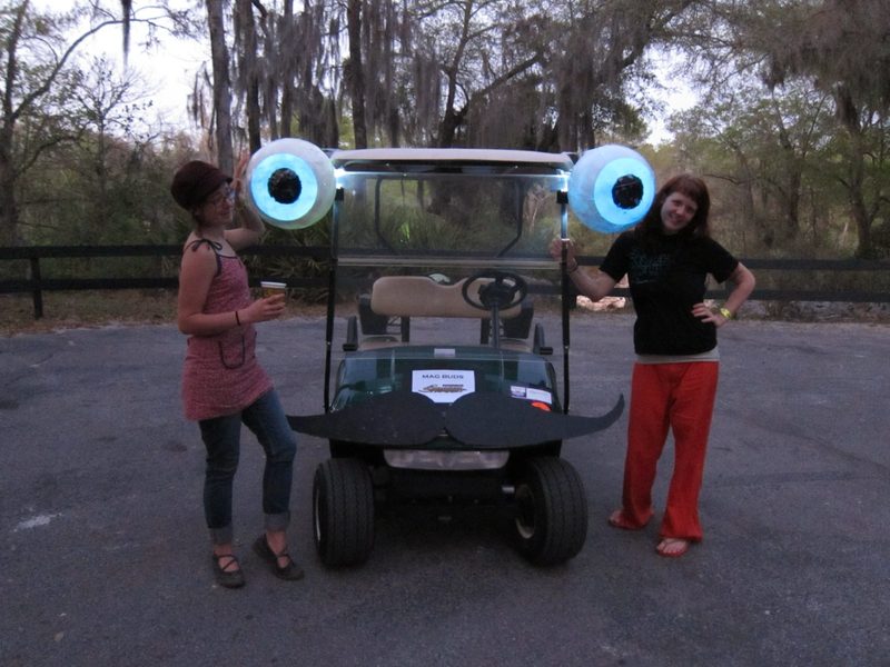 Two friends pose beside the original Zerble at dusk, each resting a hand on a giant eyeball glowing blue from the headlamp inside, above a black posterboard mustache and a MAG BUDS sign.