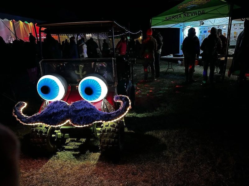 Today's Zerble at night beside a festival beer tent, its giant eyes glowing blue and the purple mustache outlined in warm lights.