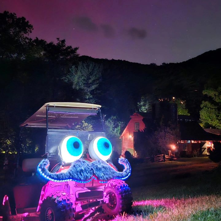 Zerble glowing at night at LEAF under a pink and purple northern-lights sky, eyes lit bright, mustache trimmed in blue lights, with a red barn in the background.