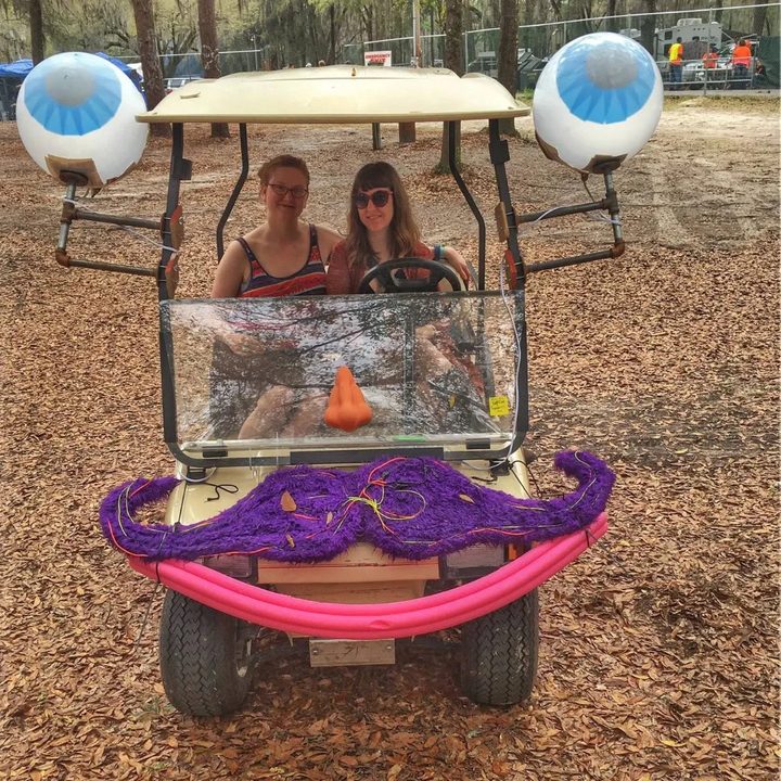 Two friends sit in Zerble at Suwannee SpringFest 2016: a cream golf cart with eyeballs on pipe stalks, an orange nose on the windshield, a fuzzy purple mustache laced with EL wire, and a pink pool-noodle smile.